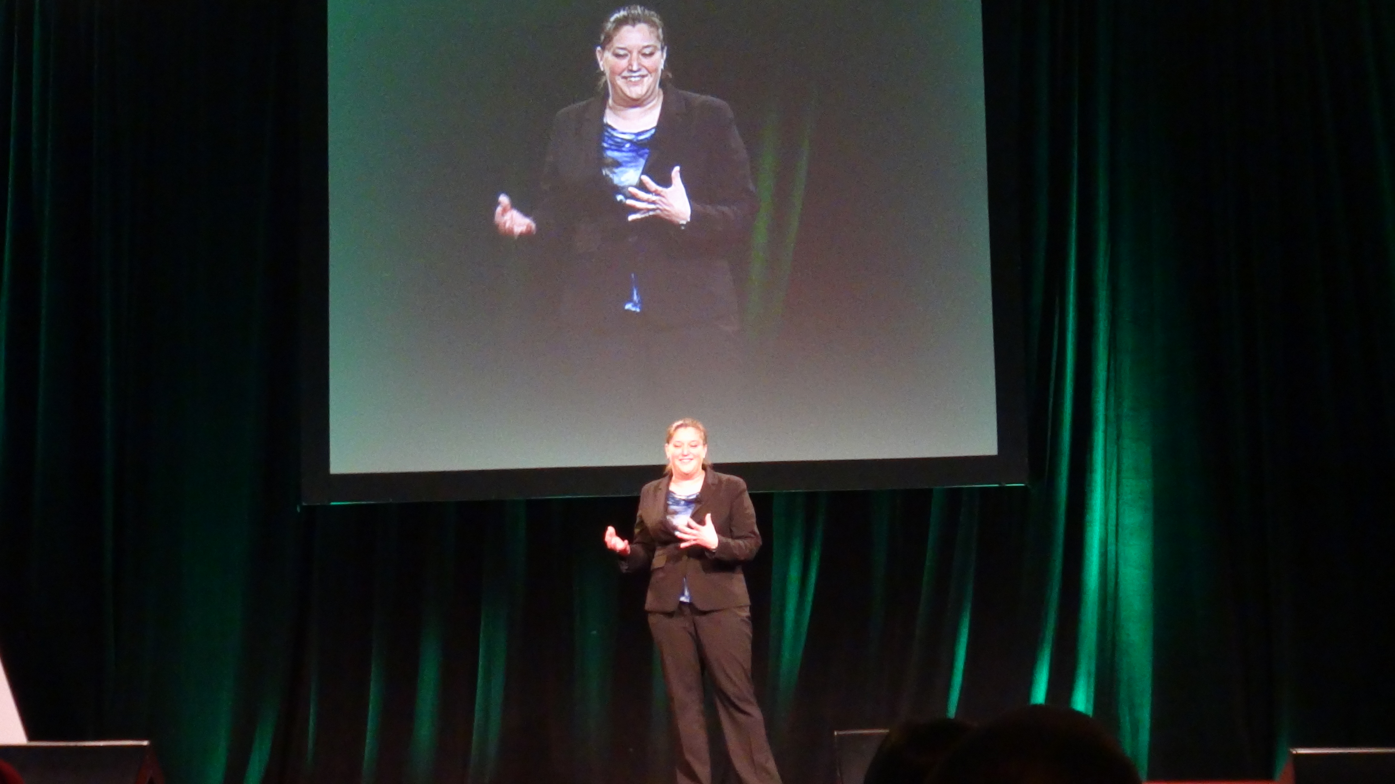 Erika a woman in a suit presenting on stage with a screen behind her and audience