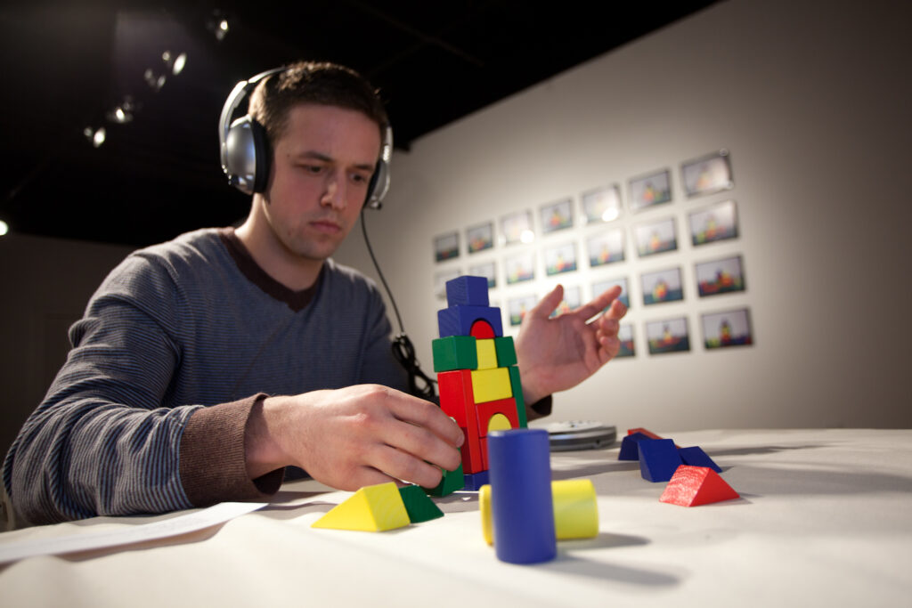 man sitting with headphones on building with multicolored blocks in front of a grid of photographs of buildings
