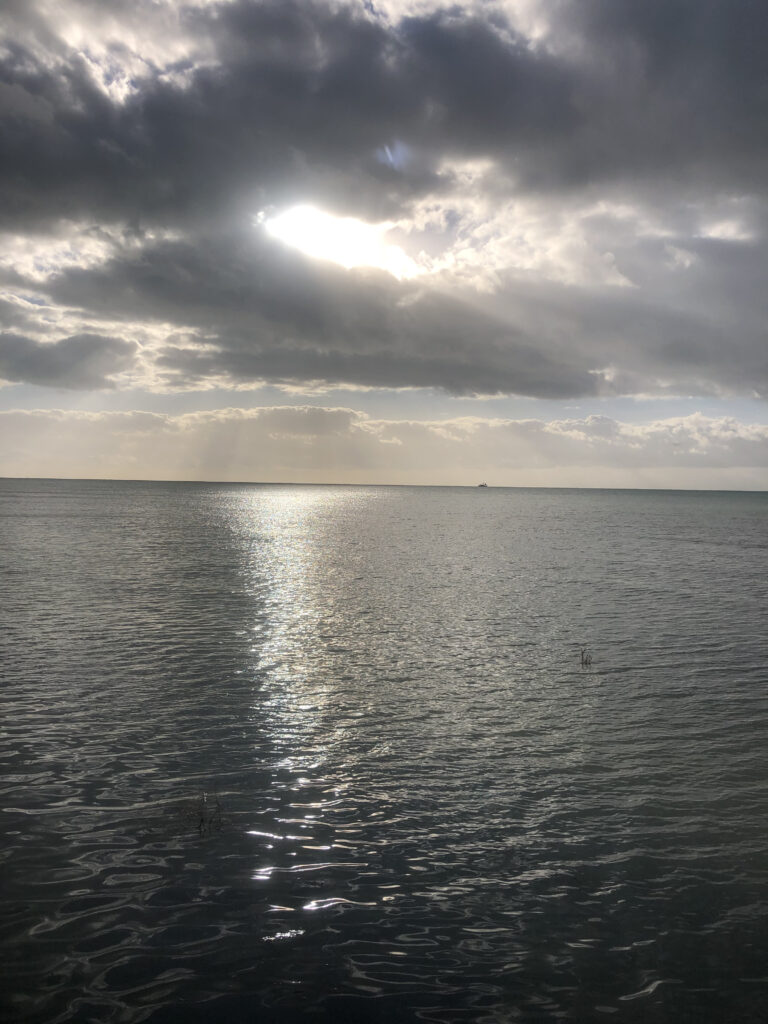 Photo by Erika Heffernan Day 269 at Smather’s Beach Boardwalk Key West. Vertical, Sun peaking through the gray clouds over a sparkling ocean