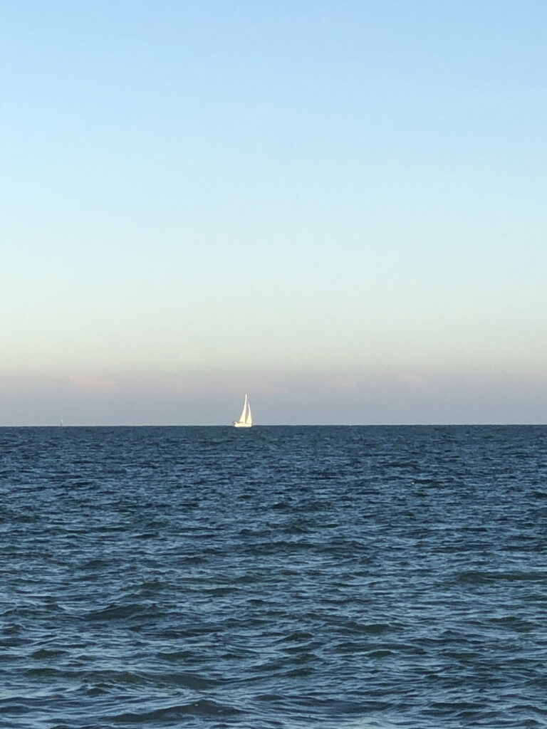 Photo by Erika Heffernan Day 271 at Smather’s Beach Boardwalk Key West. Vertical, gradations of a blue and pink sunrise sky with a white sailboat sailing in the center of the deep blue ocean with waves
