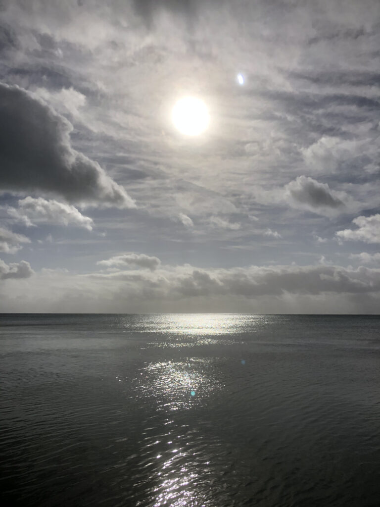 Photo by Erika Heffernan Day 286 934 AM at Smather’s Beach Boardwalk Key West. Vertical, Gray clouds in sky with sun ball bright in center over a shimmering gray ocean