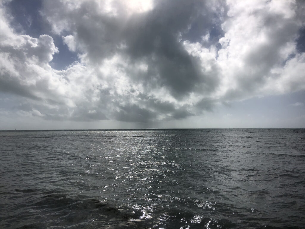 Photo by Erika Heffernan Day 293 at Smather’s Beach Boardwalk Key West. Horizontal, Sun shining through clouds over the ocean rain lines in the distance