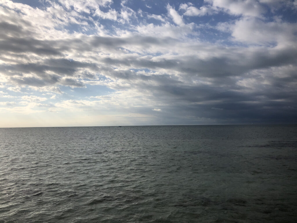 Photo by Erika Heffernan Day 299 853 AM at Smather’s Beach Boardwalk Key West. Horizontal, gray ocean with a field of clouds in the blue sky 