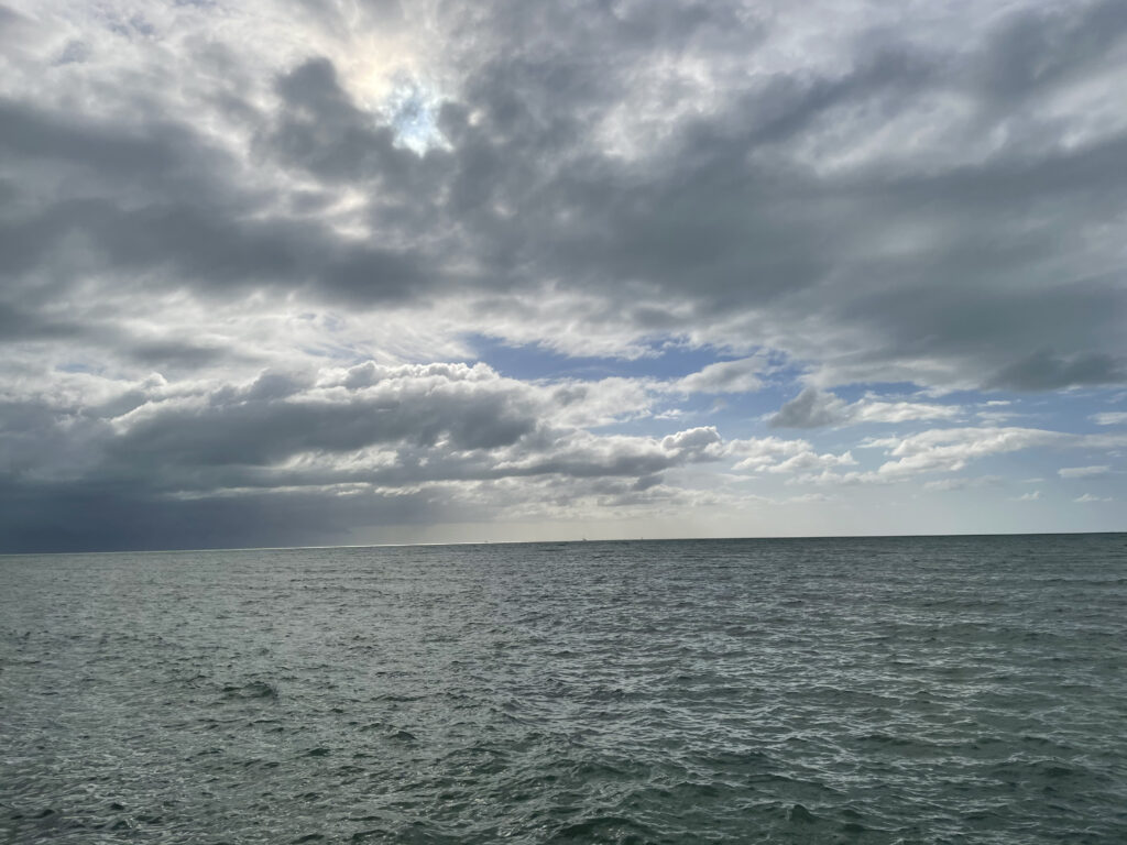 Photo by Erika Heffernan Day 323 at Smather’s Beach Boardwalk Key West. Horizontal, Blue green gray ocean with stormy clouds overhead and a rain storm on the left side moves to clear skies on the right side
