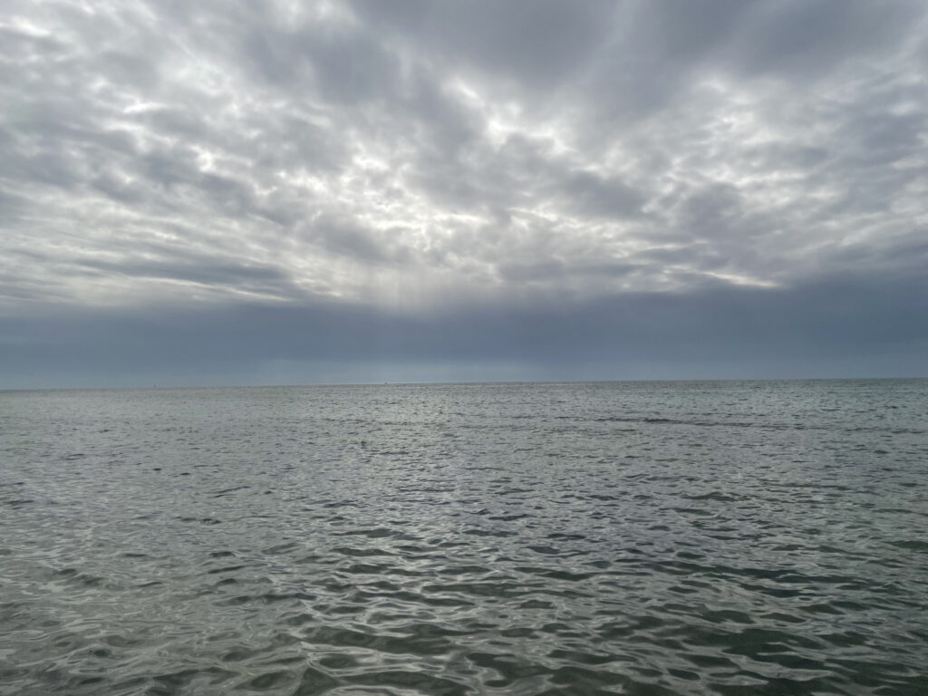 Photo by Erika Heffernan Day 329 10:02 AM at Smather’s Beach Boardwalk Key West. Horizontal cloud covered gray blue sky over a rippled gray green ocean.