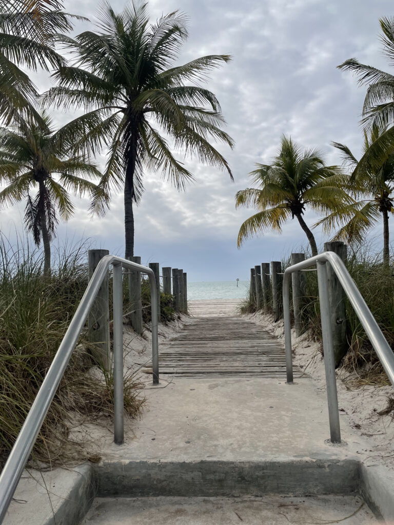 Photo by Erika Heffernan Day 329 9:53 AM at Smather’s Beach Boardwalk Key West. Vertical, Sandy stairway to to beach with wood walkway. Palm trees line the walkway with the sand beach and ocean in the distance. Dense clouds overhead with a baby blue sky.