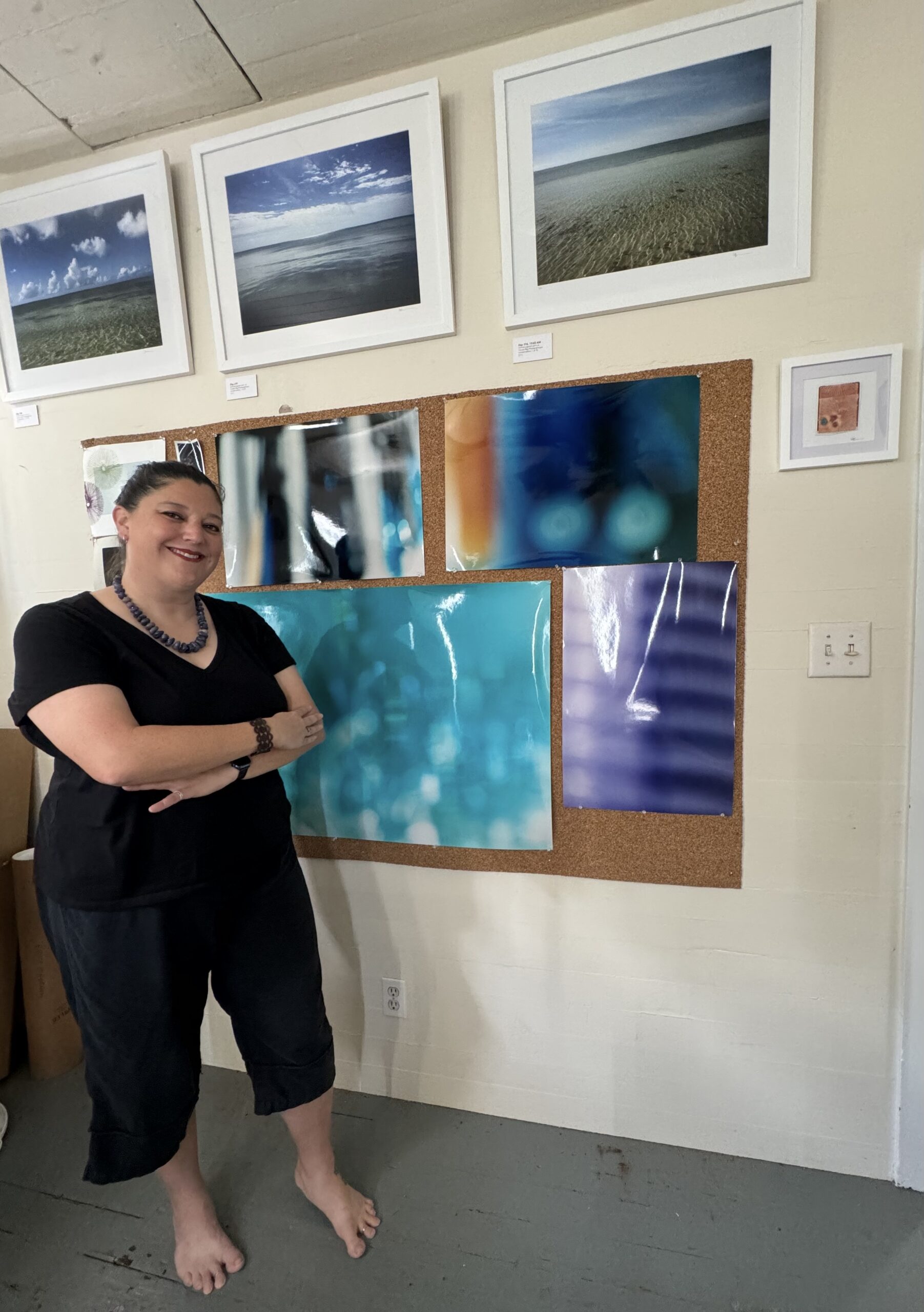 Artist Erika Heffernan standing in her Studio. woman with dark hair dressed in black with bare feet. Surounded by art to purchase.